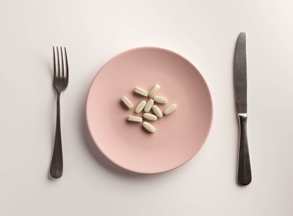 A pink plate with several vitamin pills, placed between a fork and knife on a white table.